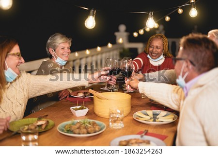 Happy multiracial senior people enjoy dinner and cheering with wine while wearing surgical face mask under chin - Coronavirus lifestyle