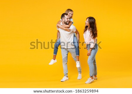 Full length portrait of laughing young parents mom dad with child kid daughter teen girl in t-shirts giving piggyback ride to joyful sit on back isolated on yellow background. Family day concept