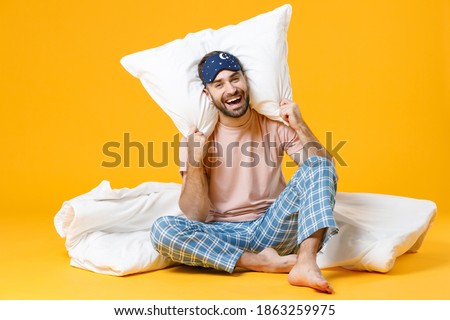 Full length of laughing young bearded man in pajamas home wear sleep mask sitting with pillow blanket isolated on bright yellow colour background studio portrait. Relax good mood lifestyle concept Royalty-Free Stock Photo #1863259975