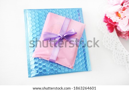 Gift wrapped with pink tissue paper and purple satin bow. Blue bubble envelope. Light background with pink flowers.
