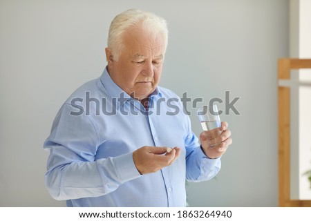 Doubtful treatment. Unsure senior man holding medical pill and glass of water thinking whether the medication is worth it, deciding whether to take it or not. Healthcare and therapy options concept Royalty-Free Stock Photo #1863264940