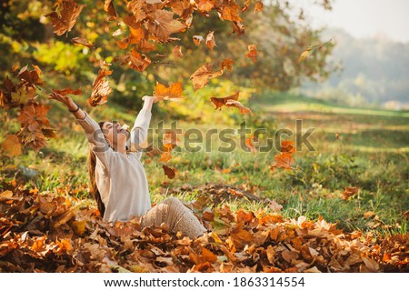 Autumn leaves falling on happy young woman in forest. The colors and mood of autumn. Beautiful young woman throwing leaves in a park. Fall concept. Happy smiling girl with natural red hair