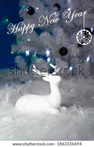 White deer in the Christmas decor. The blurred background is a white Christmas tree with openwork Christmas balls. Happy New year 2021 text.