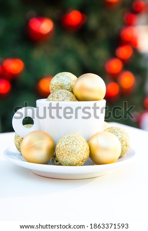 Vertical picture of coffee cup with golden Christmas balls inside and around. Coffee pair on white table angle, grey chair, new year tree with red toys in background. Winter cafe. Post card picture. 