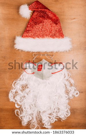 santa's hat, santa's beard and anti-battery mask on wooden background, flat lay, covid-19