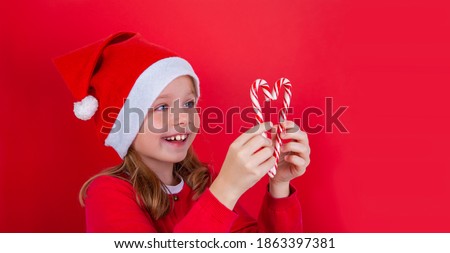 A cheerful cute girl in a new year's cap on a red background holds Christmas candy canes in her hands.