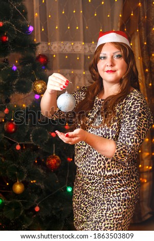 A woman in a Santa hat by a Christmas tree with a decoration ball in her hands.