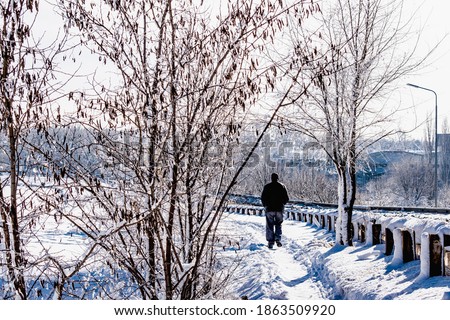 A man in warm winter clothes of black color, in trousers and jacket is walking on a snow-covered sidewalk. Around the frosty Sunny winter and a lot of blinding snow on the trees and roads. Bright blue