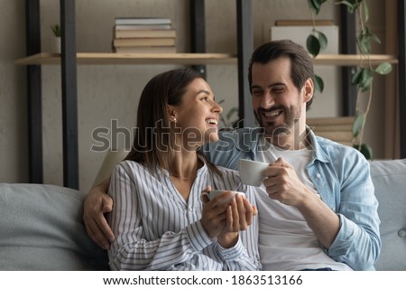 Smiling young Caucasian man and woman sit rest on sofa in living room drinking tea or coffee together. Happy millennial couple or spouses relax on couch enjoy family lazy weekend. Relaxation concept. Royalty-Free Stock Photo #1863513166