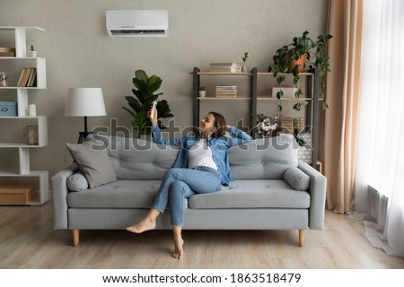 Happy young Caucasian woman relax on couch in living room turn on air conditioner with remote controller. Smiling female rest on sofa at home breathe fresh air from ac electronic condition device. Royalty-Free Stock Photo #1863518479