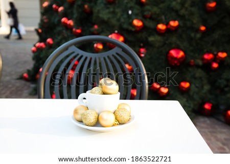 Shopping village cafe. Coffee cup with golden Christmas balls inside and around. Coffee pair on white table angle, grey chair, new year tree with red toys in background. Winter cafe. Post card picture