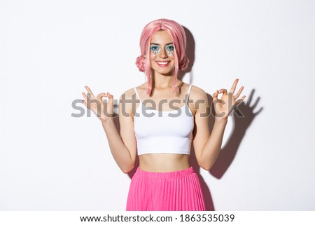 Portrait of smiling beautiful girl with pink anime wig and bright makeup, showing okay signs with satisfied expression, recommend something for halloween, standing over white background