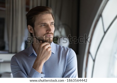 Thoughtful serious young man leader ceo executive looking at office window at workday visualizing business opportunities, dreaming of new achievements, creating personal ambitious strategy of success