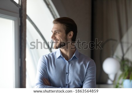 Successful smiling young man corporate employee boss team leader satisfied with professional achievements career growth standing close to window looking forward creating business vision strategy plan Royalty-Free Stock Photo #1863564859