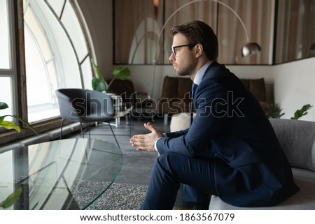 Serious thoughtful millennial male applicant candidate on vacant place expecting for appointment with hr recruiter sitting on sofa in modern office area feeling nervous worried before job interview