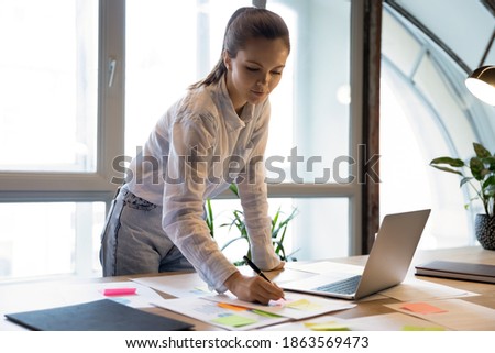 Thoughtful focused young businesswoman developing financial project on office table, preparing accounting report using computer, editing data in paper draft document adding information from internet