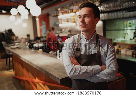 a young man of European appearance is waiting for new satisfied customers, staff is standing at the bar with his arms crossed. a Barista, employee of the waiter in restaurant cafe bistro. Royalty-Free Stock Photo #1863570178