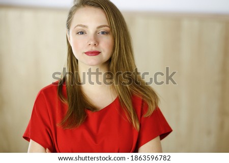 Head shot of a smart confident smiling millennial european woman standing with folded arms at home. Attractive young teenager student girl freelancer looking at camera, dressed in red