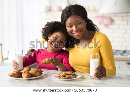 Snack Time. Happy black mother and little daughter enjoying homemade pastry and drinking milk in kitchen, having a bite with fresh baked croissants and cookies, smiling at camera, free space Royalty-Free Stock Photo #1863598870