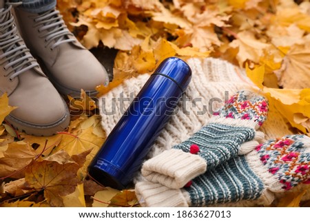 Woman standing near blue thermos, knitted scarf and mittens on fallen maple leaves outdoors, closeup