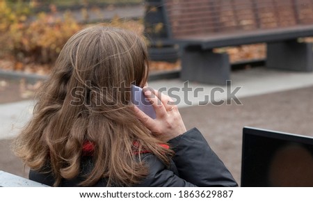 young woman sits on a park bench, speaking by the phone  and works at a computer.