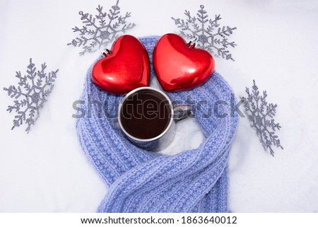 Red hearts and coffee in an old metal mug in the snow wrapped in a hand-knitted lilac scarf. New year's composition for Christmas 2021 with Christmas toys in the form of snowflakes.