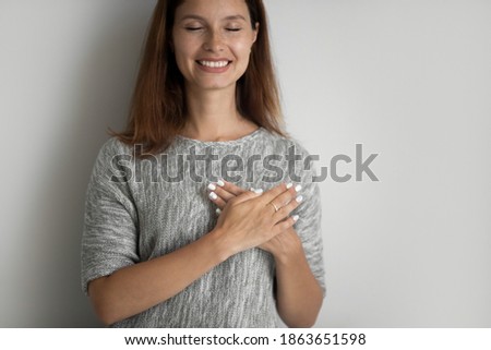 Smiling young woman holding folded hands on chest, feeling thankful showing gratitude sign isolated on grey white studio background, copy space for charity faith believe kindness advertising text.