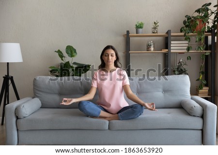 Full length peaceful young arab indian multiracial woman relaxing in lotus pose on comfortable couch, enjoying practicing yoga breathing exercises alone at home, reducing stress, healthcare concept. Royalty-Free Stock Photo #1863653920