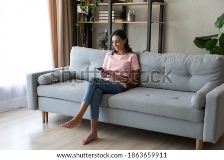 Full length happy relaxed barefoot woman sitting on cozy sofa, involved in distant mobile communication, web surfing information, shopping in online store, using telephone applications at home.