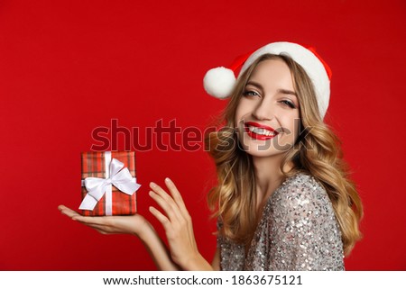Happy young woman wearing Santa hat with Christmas gift on red background