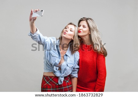 Female friends sending kiss while taking selfie. Portrait of two attractive women in stylish clothes standing, using smartphone to take funny photo. indoor studio shot isolated on gray background