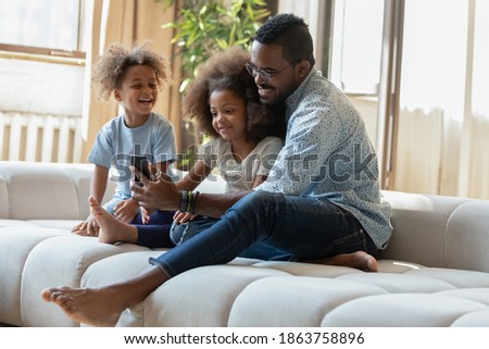 Funny movie. Caring millennial african american foster father and two little adopted kids black brother and sister having fun at home sitting on sofa close together watching cute cartoons on cellphone
