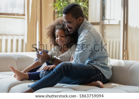 Let us call mommy. Happy african black family caring young father and smiling preteen daughter relaxing on couch hugging looking on phone screen making answering videocall, watching funny photo online