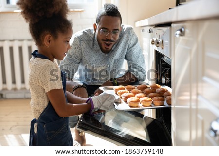 Wow, looks great! Excited caring millennial african dad or grown elder brother watching concentrated small black daughter or younger preteen sister taking pan with self baked tasty muffins out of oven Royalty-Free Stock Photo #1863759148