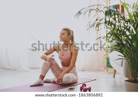 Smiling beautiful attractive young strong sporty fitness woman wearing pink tracksuit doing yoga exercises sitting holding bottle of water stretching on mat floor at home indoor