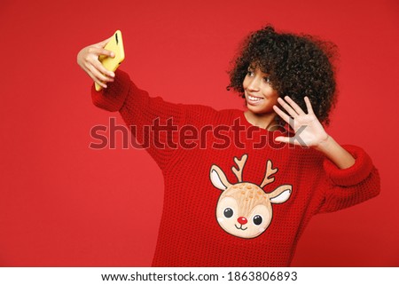 Little african curly kid girl 12-13 years old wear knitted cozy deer Christmas sweater using mobile phone doing selfie isolated on red background children studio portrait. Childhood lifestyle concept