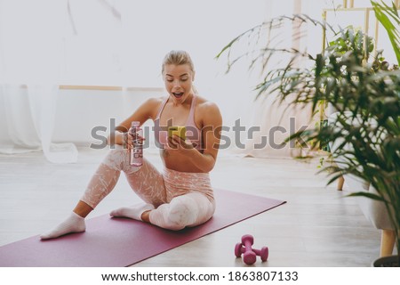 Surprised young strong sporty fitness woman wearing pink tracksuit doing yoga exercises sitting hold bottle of water using mobile cell phone typing sms message stretching on mat floor at home indoor
