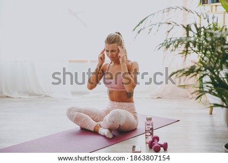 Displeased tired young strong sporty fitness woman wearing pink tracksuit doing yoga exercises sitting put hands on head keeping eyes closed having headache stretching on mat floor at home indoor