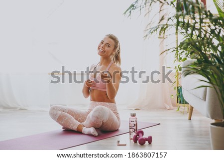 Smiling cheerful attractive beautiful young strong sporty fitness woman wearing pink tracksuit doing yoga exercises sitting pointing index fingers on camera stretching on mat floor at home indoor