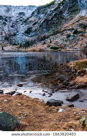 The mountain pond is partially frozen. Above, rocky mountains covered with snow and green bushes. Stones and yellow grass at the mouth of the pond.