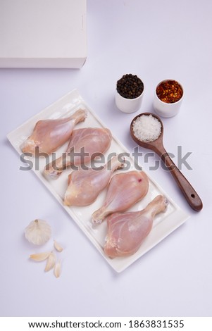 Raw chicken drum sticks arranged on white table ware with black pepper,chilli flakes,salt and garlic with delivery box on white colour background, top view.