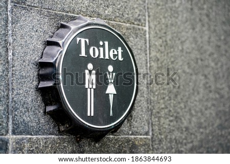 A toilet sign on a black beer bottle cap attached on cement wall texture with copy space