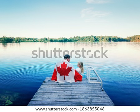 Father and daughter wrapped in large Canadian flag sitting on wooden pier by lake. Canada Day celebration outdoor. Dad and child sitting together on 1 of July celebrating national Canada Day. Royalty-Free Stock Photo #1863855046