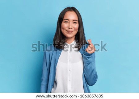 Lovely brunette Asian woman with rouge cheeks show mini heart gesture makes korean like sign wears white shirt and blue jumper expresses love has gentle smile poses indoor. Body language concept