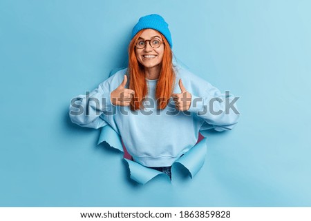 Cheerful redhead European woman makes thumb up gesture makes excellent sign approves something smiles broadly wears hat and sweater breaks through paper background gives advice says I like it