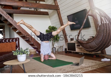 Full length shot of a sporty middle aged man standing in headstand pose while exercising at home and watching online video tutorial on laptop