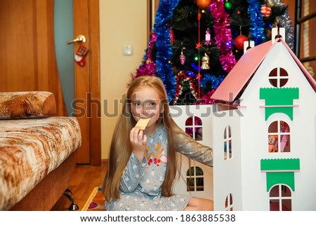 A little girl in a gray dress eats chips near a Dollhouse and a decorated Christmas tree.