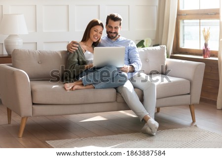 Happy young Caucasian couple renters relax on sofa in living room watch video on laptop together. Smiling man and woman tenants rest on comfortable couch at home use modern computer gadget. Royalty-Free Stock Photo #1863887584