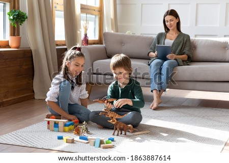 Happy two little siblings children relax on warm floor in cozy living room play toys together, mom relax on couch with tablet. Smiling kids engaged in game, mother rest using pad device at home. Royalty-Free Stock Photo #1863887614