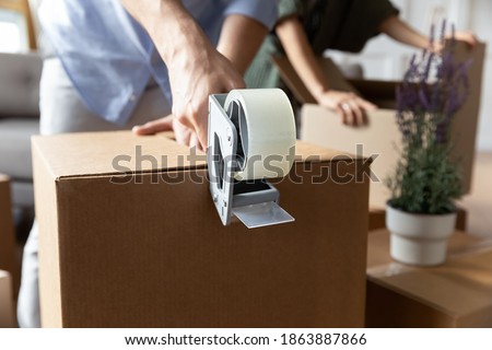Crop close up of man renter tenant pack box with personal belonging with adhesive scotch on moving day. Husband wrap package with tape dispenser, relocate move to new home. Delivery concept. Royalty-Free Stock Photo #1863887866